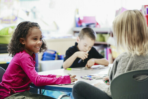 Two children sit at a table with papers and pens. A teacher sits as well.